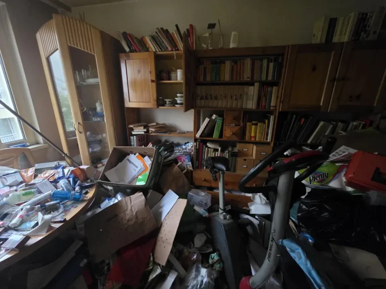Messie Entrümpelung 1030 Wien Landstraße – Wohnraum vor der Räumung Messie Entrümpelung 1030 Wien Landstraße vorher: Wohnraum mit Bücherregalen, überladenem Tisch, Kartons und Heimtrainer
